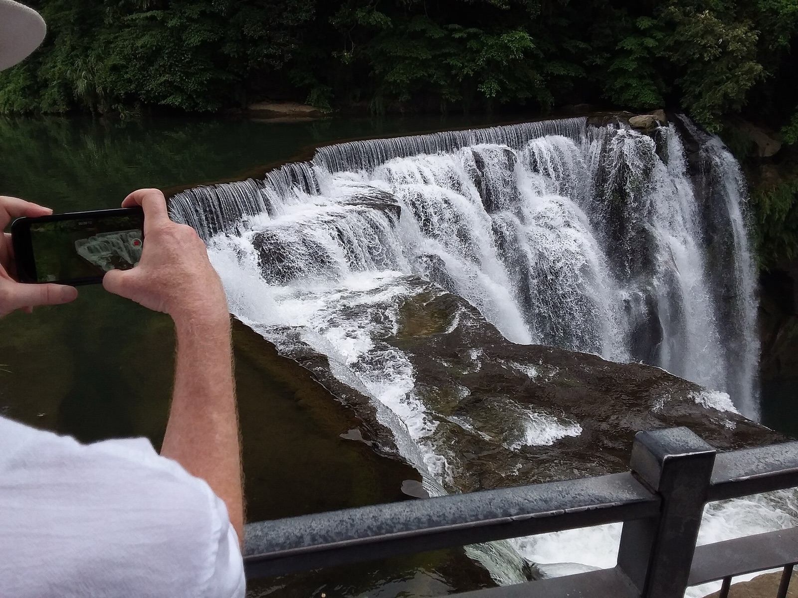 A person holds an iPhone to film a waterfall from a railing, with the phone's screen showing the view.
