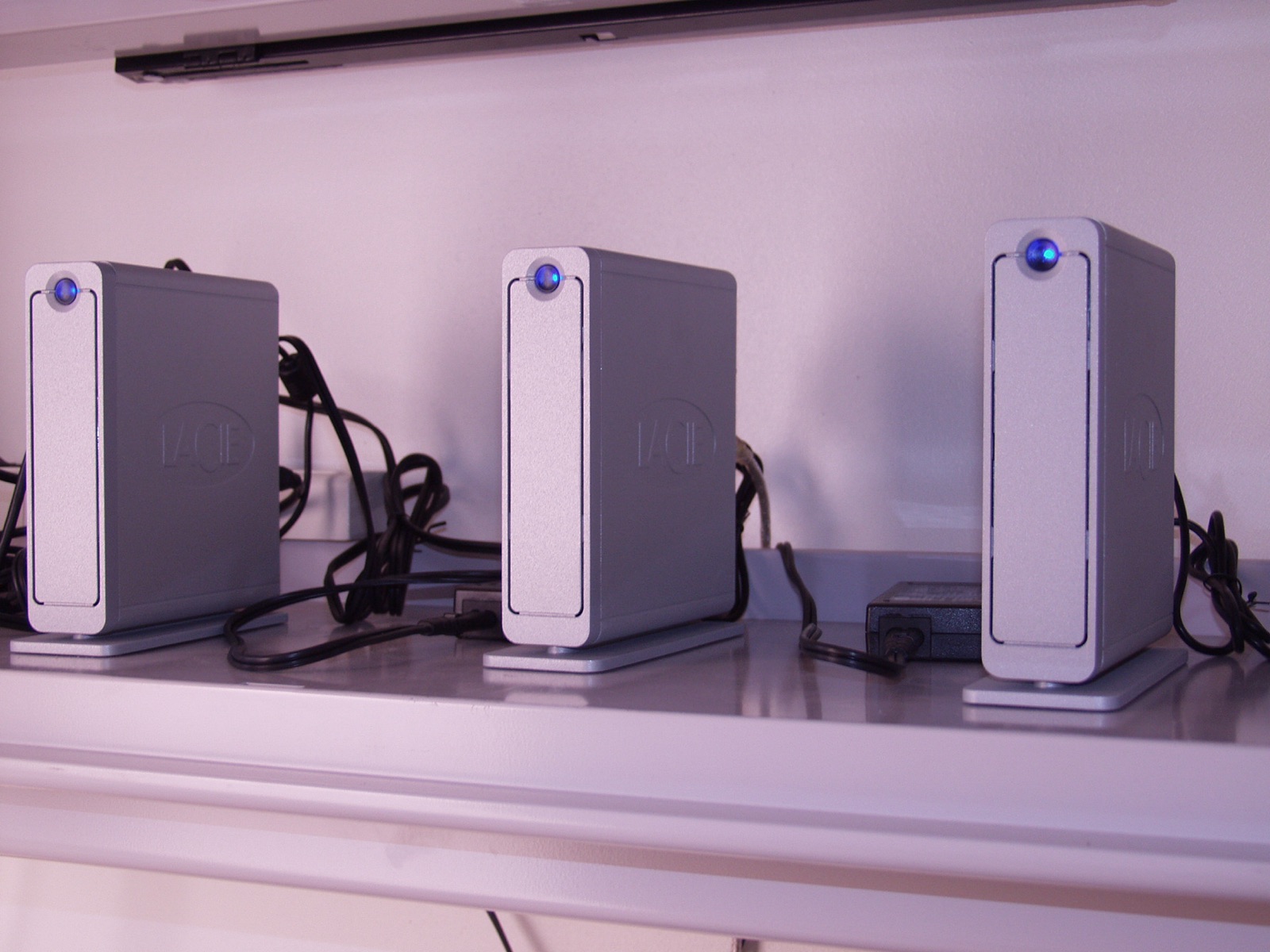 Three silver external hard drives in a row on a shelf, each with a small blue light.